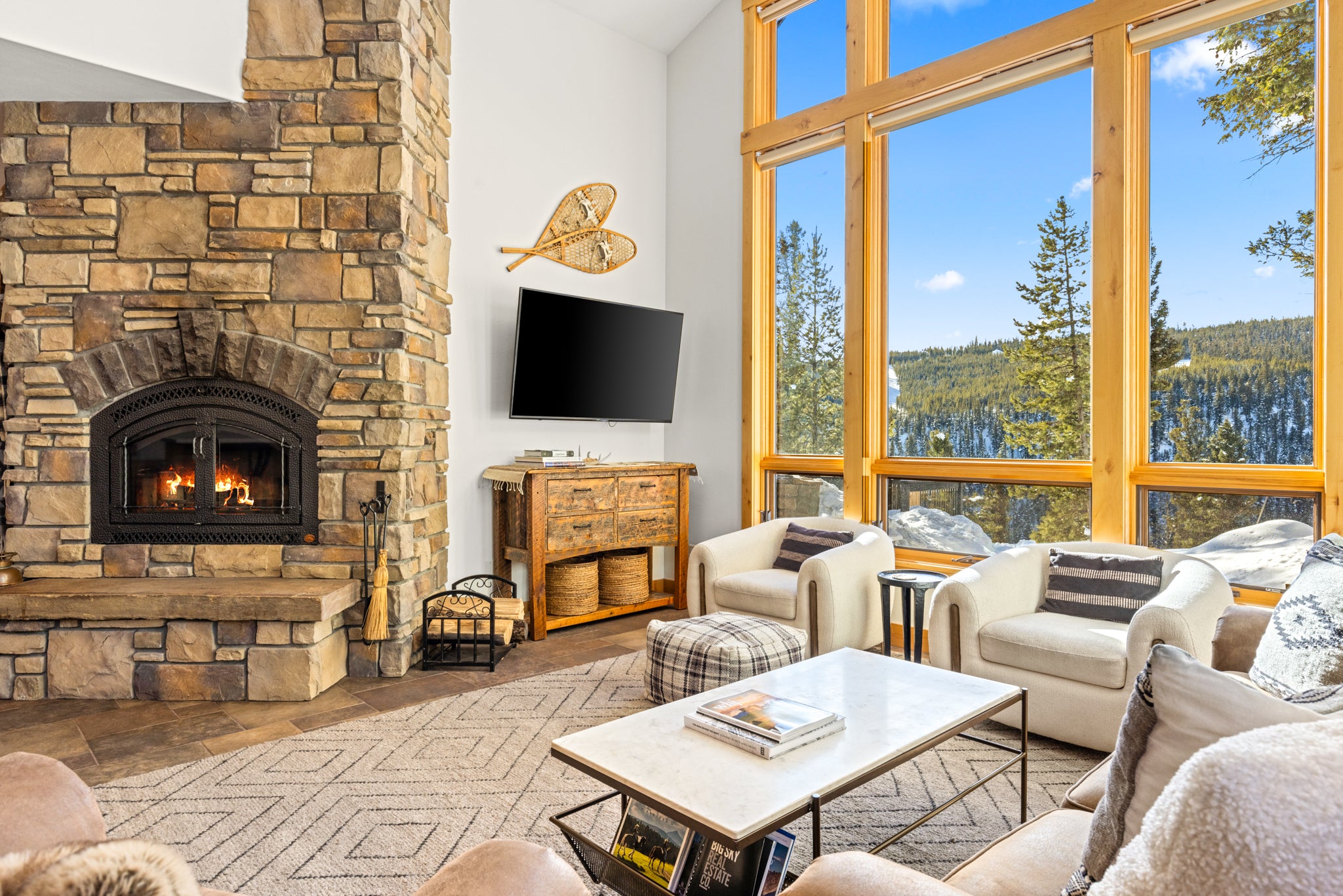 Open living room with tall windows and white furniture with a stone fireplace and TV in Big Sky, Montana
